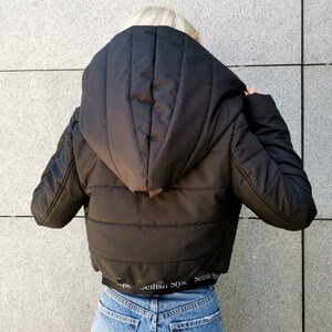 May include: Black hooded puffer jacket with a cropped hem. The jacket has a quilted design and features the text "Selfish Style" repeated around the bottom hem.