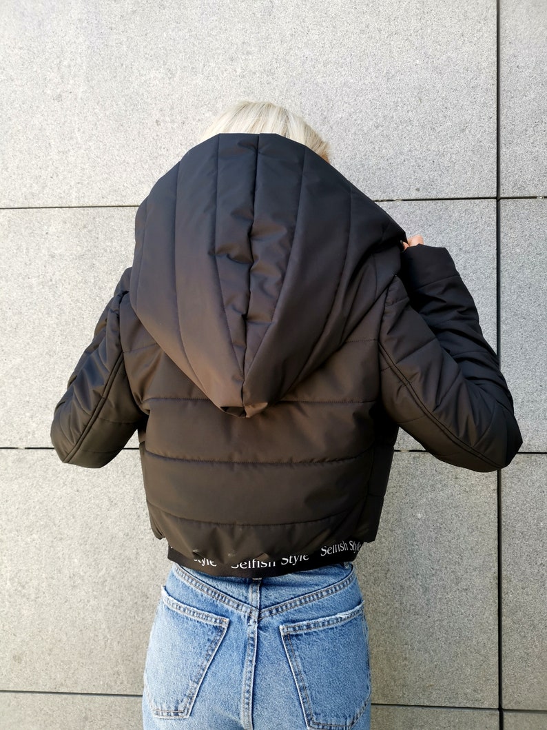 May include: Black hooded puffer jacket with a cropped hem. The jacket has a quilted design and features the text "Selfish Style" repeated around the bottom hem.