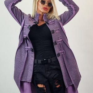 Pode incluir: Um casaco comprido roxo com gola alta e fechos decorativos. O casaco tem acabamentos roxos contrastantes e &eacute; combinado com jeans pretos rasgados e um top preto. O look &eacute; completado com &oacute;culos de sol e um cinto preto.