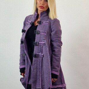 Pode incluir: Um casaco comprido roxo com fivelas e acabamentos pretos. O casaco tem um aspeto texturizado e uma gola alta. A modelo est&aacute; a usar cal&ccedil;as pretas. O casaco &eacute; uma pe&ccedil;a de roupa exterior elegante.