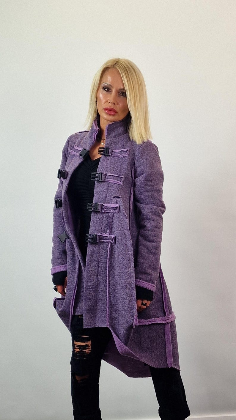 Pode incluir: Um casaco comprido roxo com fivelas e acabamentos pretos. O casaco tem um aspeto texturizado e uma gola alta. A modelo est&aacute; a usar cal&ccedil;as pretas. O casaco &eacute; uma pe&ccedil;a de roupa exterior elegante.