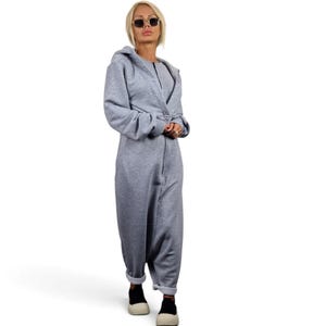 May include: A light gray hooded jumpsuit featuring long sleeves, a front zipper closure, and a relaxed fit. The garment is constructed from a soft, jersey-like fabric with ribbed cuffs at the ankles and wrists, providing a casual and comfortable silhouette.