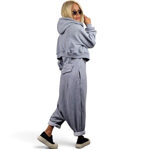 May include: A light gray hooded jumpsuit featuring a cropped hoodie top with long sleeves, a fitted waist, and loose-fitting trousers with side pockets. The garment is made of a soft jersey fabric and is paired with black platform sneakers.