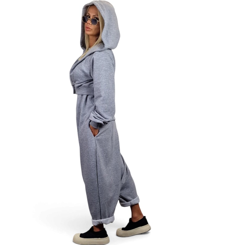 May include: A light gray hooded jumpsuit featuring long sleeves, a front zipper closure, and side pockets. The garment has a relaxed fit with cuffed ankles and is paired with black slip-on shoes with thick, textured beige rubber soles.