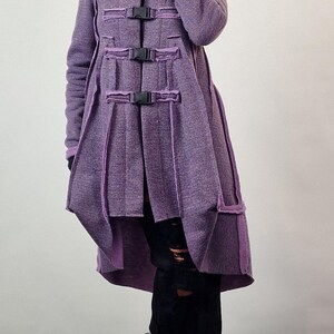 Pode incluir: Um casaco comprido roxo com fivelas pretas e um design &uacute;nico. O casaco tem uma gola alta e uma saia rodada. A pessoa est&aacute; usando cal&ccedil;as pretas e sapatos pretos.