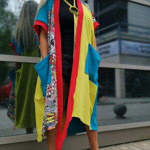 Może przedstawiać: Kobieta w kolorowej kurtce w stylu kimona w kolorze czerwonym, ż&oacute;łtym i niebieskim. Kurtka ma graficzny nadruk wewnątrz i długi, płynny kr&oacute;j. Ma na sobie czarne spodenki i czarno-białe szachownice.