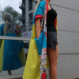 Może przedstawiać: Kobieta w długim, kolorowym kardiganie z wieloma wzorami, czarnym topie na ramiączkach i dżinsowych szortach. Ma na sobie czarno-białe sznurowane buty sportowe.