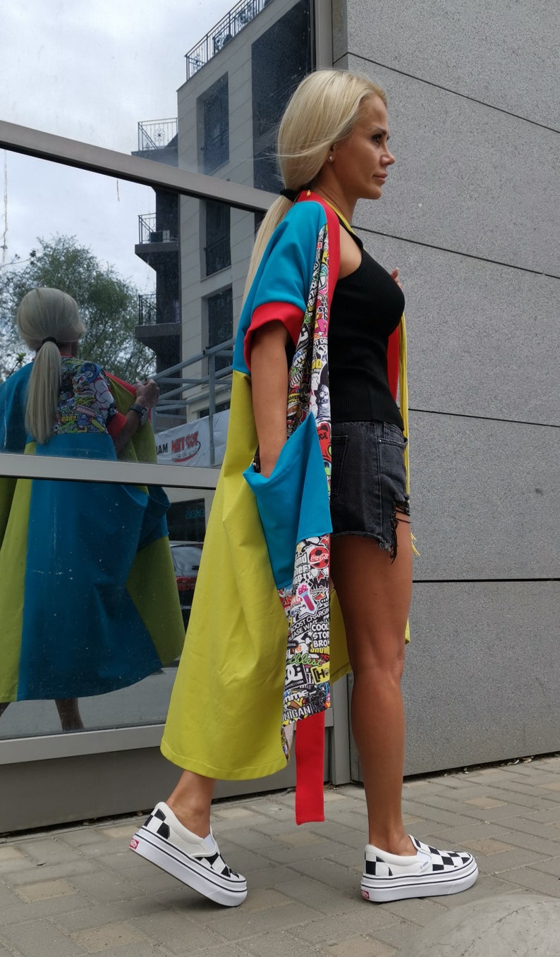 Może przedstawiać: Kobieta w długim, kolorowym kardiganie z wieloma wzorami, czarnym topie na ramiączkach i dżinsowych szortach. Ma na sobie czarno-białe sznurowane buty sportowe.