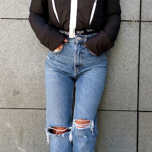 May include: A black cropped puffer jacket with white accents and a zip closure. The jacket has a hood and the text "Style Sense" is visible on the bottom of the jacket. The model is wearing blue jeans with distressed details and black fringe boots.