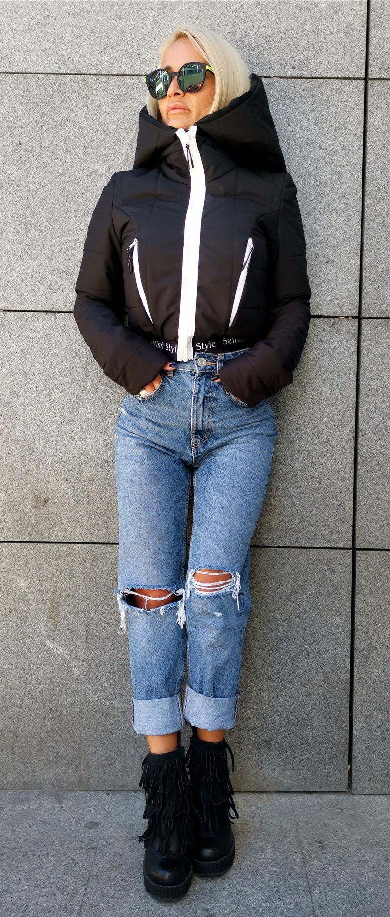 May include: A black cropped puffer jacket with white accents and a zip closure. The jacket has a hood and the text "Style Sense" is visible on the bottom of the jacket. The model is wearing blue jeans with distressed details and black fringe boots.