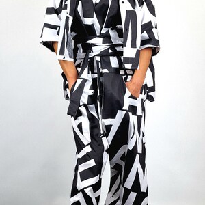 May include: A black and white patterned jumpsuit with a black belt and a black and white patterned jacket. The model is wearing black sunglasses and a black headband.