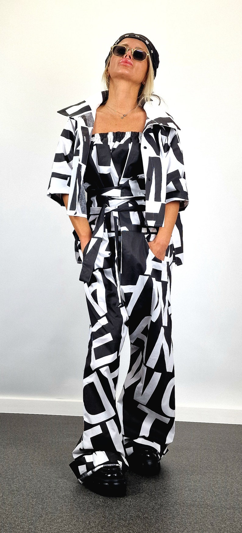 May include: A black and white patterned jumpsuit with a black belt and a black and white patterned jacket. The model is wearing black sunglasses and a black headband.