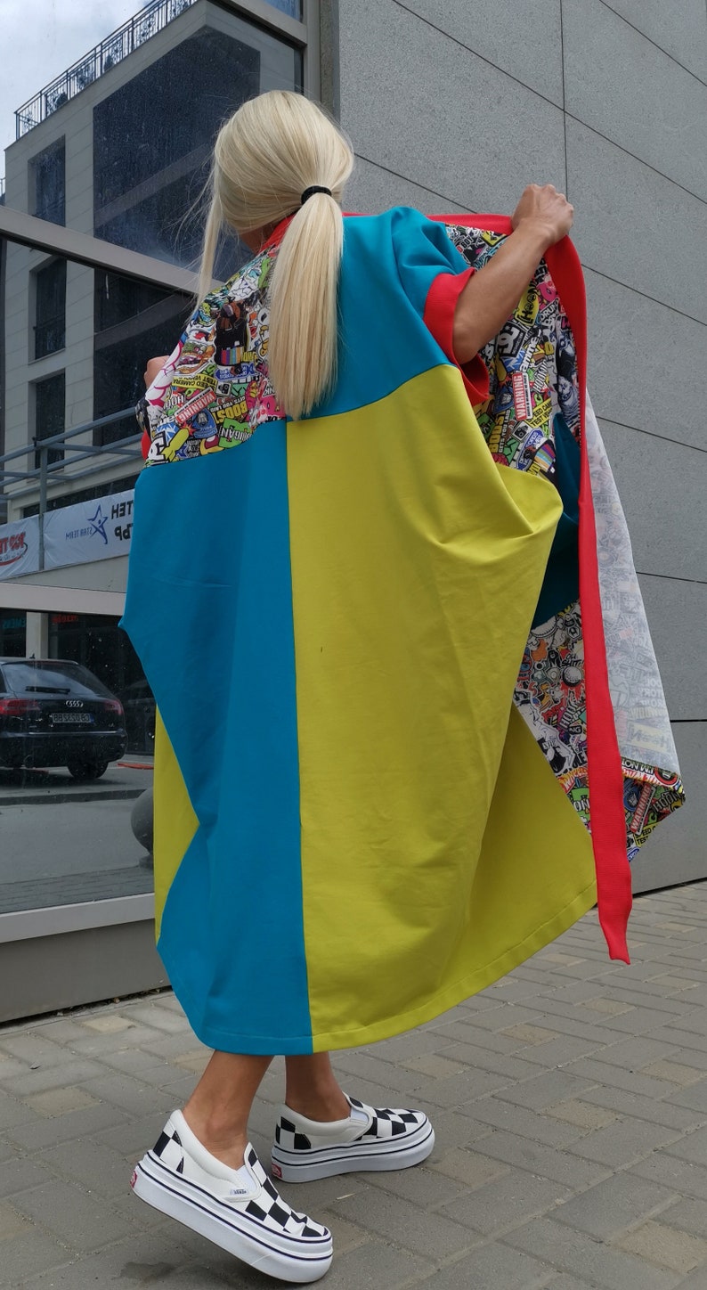 Może przedstawiać: Kobieta w długiej, kolorowej tunice w niebiesko-ż&oacute;łto-czerwonej kolorystyce. Tunika ma graficzny nadruk z naklejkami i logo. Ma na sobie czarno-białe sznurowane buty.