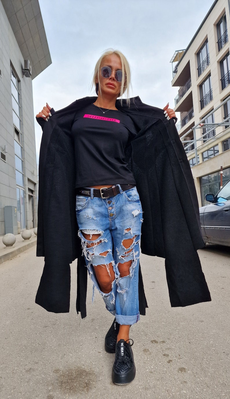 Pode incluir: Uma mulher vestindo uma camisa preta de manga comprida com o texto "Selfish" em rosa, jeans azul rasgado, cinto preto e t&ecirc;nis pretos. Ela est&aacute; usando &oacute;culos de sol e tem o cabelo puxado para tr&aacute;s.