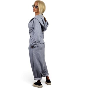 May include: A light gray hooded jumpsuit featuring long sleeves, a belted waist, and side pockets. The garment is crafted from a soft, fleece-like material with a relaxed fit, cuffed ankles, and a structured hood, providing a cohesive look.