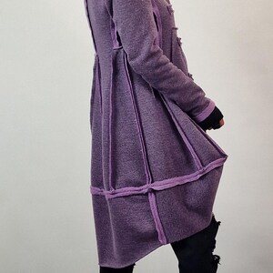 Pode incluir: Um casaco comprido roxo com um design &uacute;nico. O casaco apresenta um tecido texturizado com acabamentos e costuras roxas mais claras. &Eacute; combinado com cal&ccedil;as pretas e sapatos pretos grossos. O estilo geral &eacute; moderno e chique.