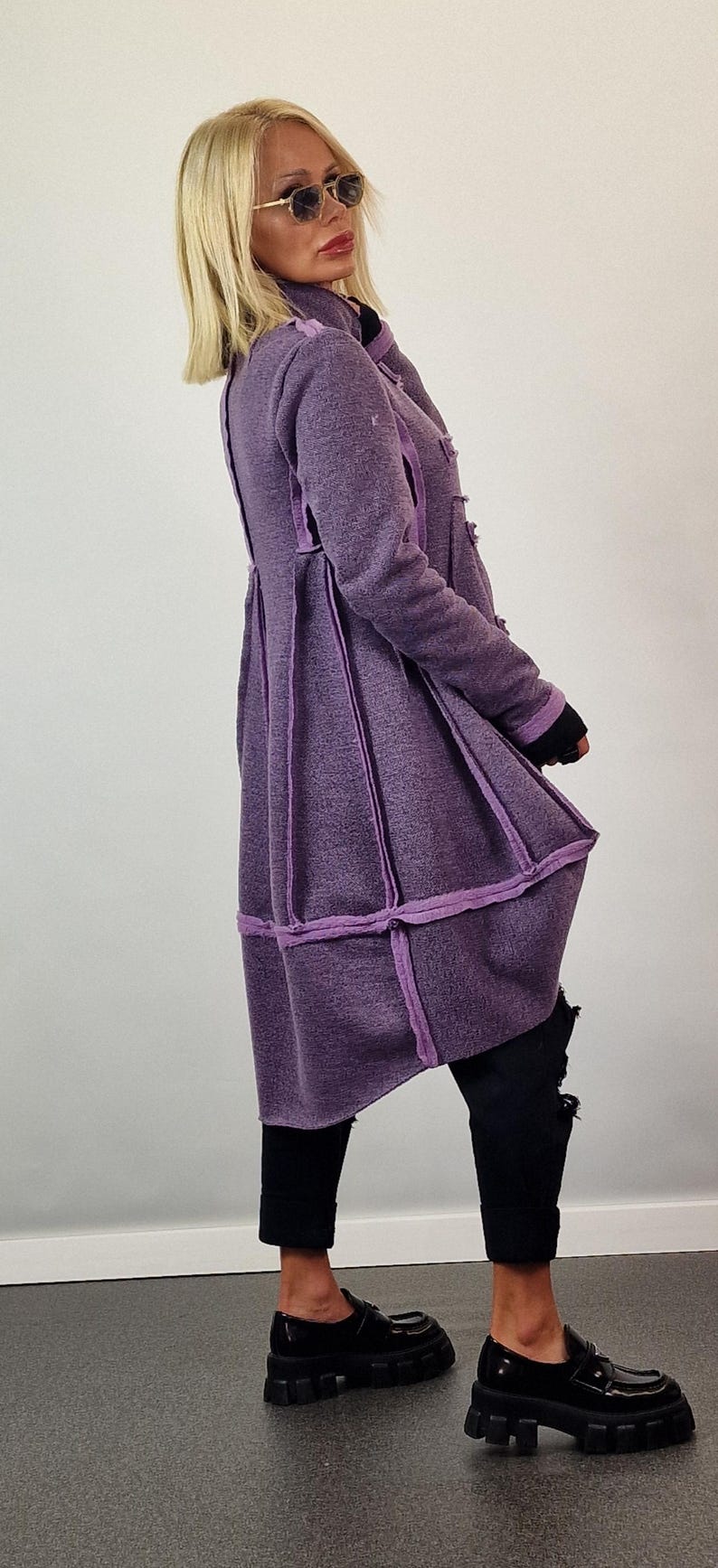 Pode incluir: Um casaco comprido roxo com um design &uacute;nico. O casaco apresenta um tecido texturizado com acabamentos e costuras roxas mais claras. &Eacute; combinado com cal&ccedil;as pretas e sapatos pretos grossos. O estilo geral &eacute; moderno e chique.
