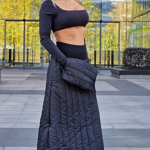 Può includere: Una donna indossa un top corto nero a maniche lunghe, leggings neri e una gonna trapuntata nera. Indossa occhiali da sole e tiene una borsa nera in mano.