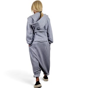 May include: A light gray hooded jumpsuit featuring long sleeves, a structured hood, a horizontal waistband detail, and a flap pocket on the back. The garment has a relaxed fit with tapered legs that end above the ankles, showcasing a clean, solid-colored design.