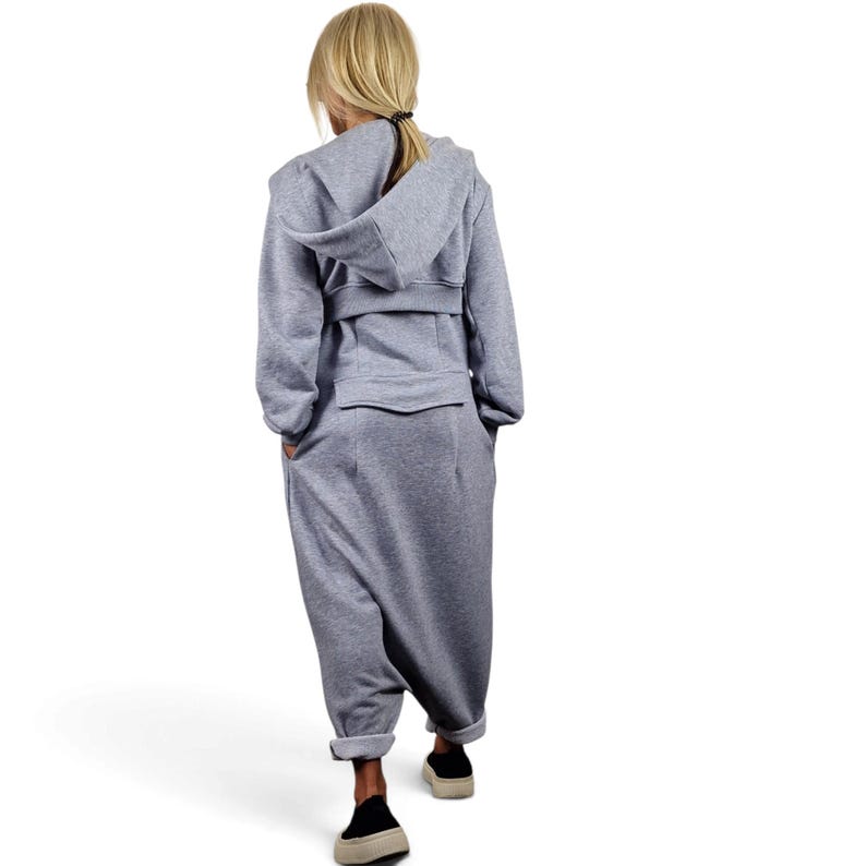 May include: A light gray hooded jumpsuit featuring long sleeves, a structured hood, a horizontal waistband detail, and a flap pocket on the back. The garment has a relaxed fit with tapered legs that end above the ankles, showcasing a clean, solid-colored design.