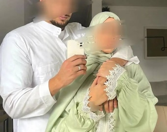 Può includere: Una coppia vestita con abiti islamici tradizionali. La donna indossa un'abaya verde chiaro con bordi in pizzo bianco e un velo abbinato. L'uomo indossa una thobe bianca. Stanno tenendo un telefono cellulare e sorridendo.