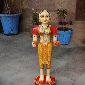May include: A hand-painted wooden figurine of a woman wearing a red and yellow sari with intricate details. The figurine is standing on a red and yellow base with a gold trim.