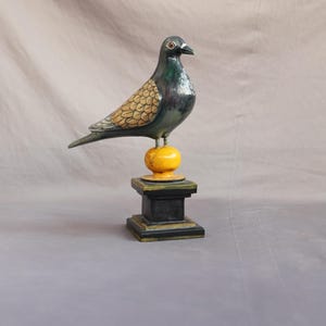 May include: A decorative wooden pigeon figurine. The bird is perched atop a yellow sphere, which is mounted on a black square pedestal. The pigeon has a dark green body, tan wings, and a black beak. The sculpture is approximately 10 inches tall.