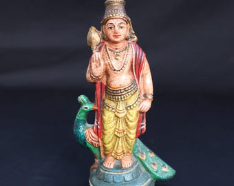 Puede incluir: Estatua pintada a mano de una deidad, con una figura con ropas amarillas y rojas, adornada con joyas y un tocado. La figura está sobre una base turquesa con un pavo real. La estatua mide unos 15 cm de altura.