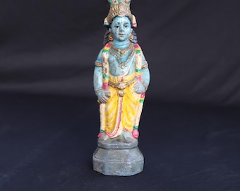 Puede incluir: Pequeña estatua pintada a mano de una deidad con piel azul, que lleva un dhoti amarillo y una corona enjoyada. La estatua tiene una expresión serena y está adornada con collares y guirnaldas. Se alza sobre un pedestal gris sobre un fondo oscuro.