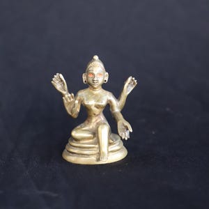 May include: A small, brass-colored statue of a seated female figure with four arms. The figure has detailed facial features, including red-colored eyes, and is seated on a tiered base. The statue is set against a dark background.