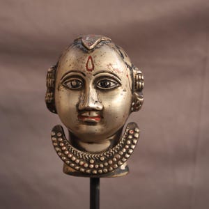 May include: A silver-coloured sculpture of a human head with detailed facial features, including dark eyes, red lips, and a red dot on the forehead. The head is adorned with decorative elements and a beaded collar.