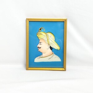 May include: A framed painting of a man in a yellow turban with a blue background. The man is wearing a white shirt and has a mustache.