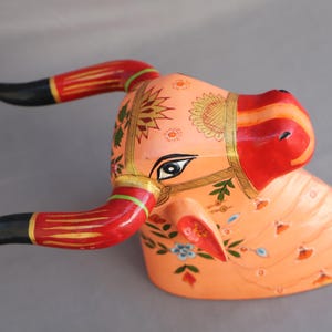 Pu&ograve; includere: Scultura di testa di toro in legno dipinta a mano con dettagli intricati. La testa del toro &egrave; dipinta in tonalit&agrave; di rosa, rosso e oro con motivi floreali. Le corna sono nere con strisce rosse e dorate.