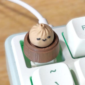 May include: A beige dumpling keycap with a smiling face sits in a brown steamer basket, placed on a white keyboard. The keycap is on the F1 key. A white cable is visible in the background.
