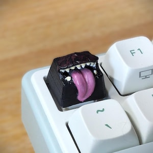 Könnte beinhalten: Eine individuelle Tastenkappe mit einem Monster-Design. Die schwarze Tastenkappe hat ein detailliertes Monstergesicht mit weißen Zähnen und einer rosa Zunge. Die Tastenkappe befindet sich auf einer weißen und hellblauen Tastatur.