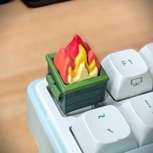 May include: A custom mechanical keyboard keycap shaped like a green dumpster with bright red and yellow flames emerging from the top. The keycap is installed on a white keyboard next to an F1 key and a key with a tilde symbol, replacing a standard key.