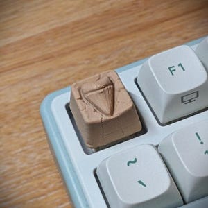 May include: A light brown, textured artisan keycap featuring a raised shark tooth design, installed on a computer keyboard next to a white F1 key with a green icon and a white key with a green tilde symbol on a light blue keyboard frame.
