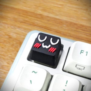 May include: A black mechanical keyboard keycap featuring a white graphic of a smiling face with closed eyes and red blush marks on the cheeks, positioned on a white keyboard next to an F1 key with a monitor icon and other standard white keys.