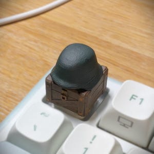 May include: A custom mechanical keyboard keycap featuring a miniature dark green helmet resting on top of a brown wooden crate. The crate has visible plank textures and a small central latch detail. The keycap is installed on a white keyboard.