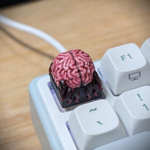 Peut inclure: Une touche de clavier mécanique personnalisée représentant une sculpture de cerveau rose texturée et détaillée sur une base noire avec des vrilles sombres et dégoulinantes, placée entre des touches blanches sur un clavier clair.