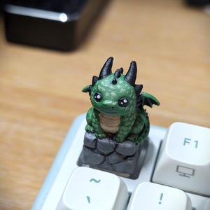 Peut inclure: Une touche de clavier mécanique personnalisée représentant un petit dragon vert avec des cornes et des ailes noires, perché sur une base grise texturée en pierre. Le dragon a un ventre clair et de grands yeux sombres, posé sur une touche blanche.