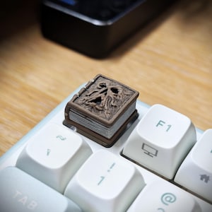 May include: A brown, square-shaped artisan keycap designed to resemble a miniature book with a sculpted, weathered skull face on the cover. The book features detailed page edges in a light grey colour, positioned on a white computer keyboard.