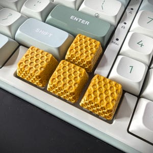 Op de afbeelding: Close-up van een computertoetsenbord met aangepaste keycaps. De keycaps zijn geel en hebben een honingraatpatroon. De toetsen van het toetsenbord zijn wit, met een lichtblauwe shift-toets en een groene enter-toets.