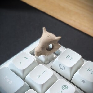 May include: A tan-coloured, skull-shaped keycap is installed on a white keyboard. The keycap is a detailed, three-dimensional design. The keyboard has white keys with light green symbols and letters.