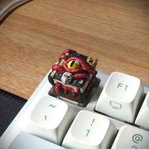 以下が含まれることがあります： ファンタジーをテーマにしたカスタムアーティザンキーキャップ。赤い質感の表紙、黄色い目、頭蓋骨が付いた小さな開いた本が描かれています。キーキャップは、薄緑色の文字が入った白いキーボードに配置されています。