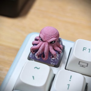 Peut inclure: Une touche de clavier mécanique personnalisée représentant une pieuvre rose sculptée avec des tentacules texturés reposant sur une base violette. La touche est installée sur un clavier blanc à côté d'une touche marquée F1 et d'une icône d'ordinateur.