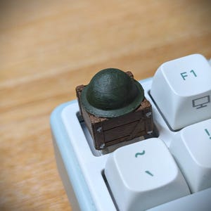 May include: A custom mechanical keyboard keycap shaped like a brown wooden crate topped with a dark olive green Brodie helmet. The keycap is installed on a white keyboard base next to a white keycap labeled with the text F1 and a monitor icon.