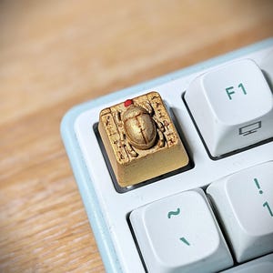Peut inclure: Une touche de clavier artisanale dorée présentant un motif de scarabée en relief avec un petit accent rouge sur la tête, entouré de symboles gravés de style hiéroglyphique, installée sur un clavier mécanique blanc à côté de la touche F1.