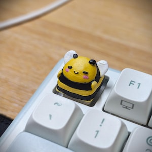 May include: A custom keyboard keycap shaped like a small, yellow and black striped bee with white wings, closed eyes, and pink cheeks, installed on a white mechanical keyboard next to the F1 key and the tilde key on a standard computer layout.
