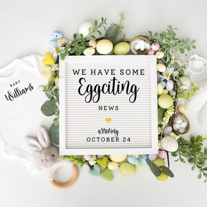 Pode incluir: Um quadro de letras branco com o texto "We have some Eggciting News arriving October 24" rodeado por uma coroa de vegetação, ovos coloridos e itens para bebês como um macacão, sapatinhos e um coelho de pelúcia.